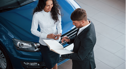 Man and woman in business attire standing by a blue car, reviewing documents.
