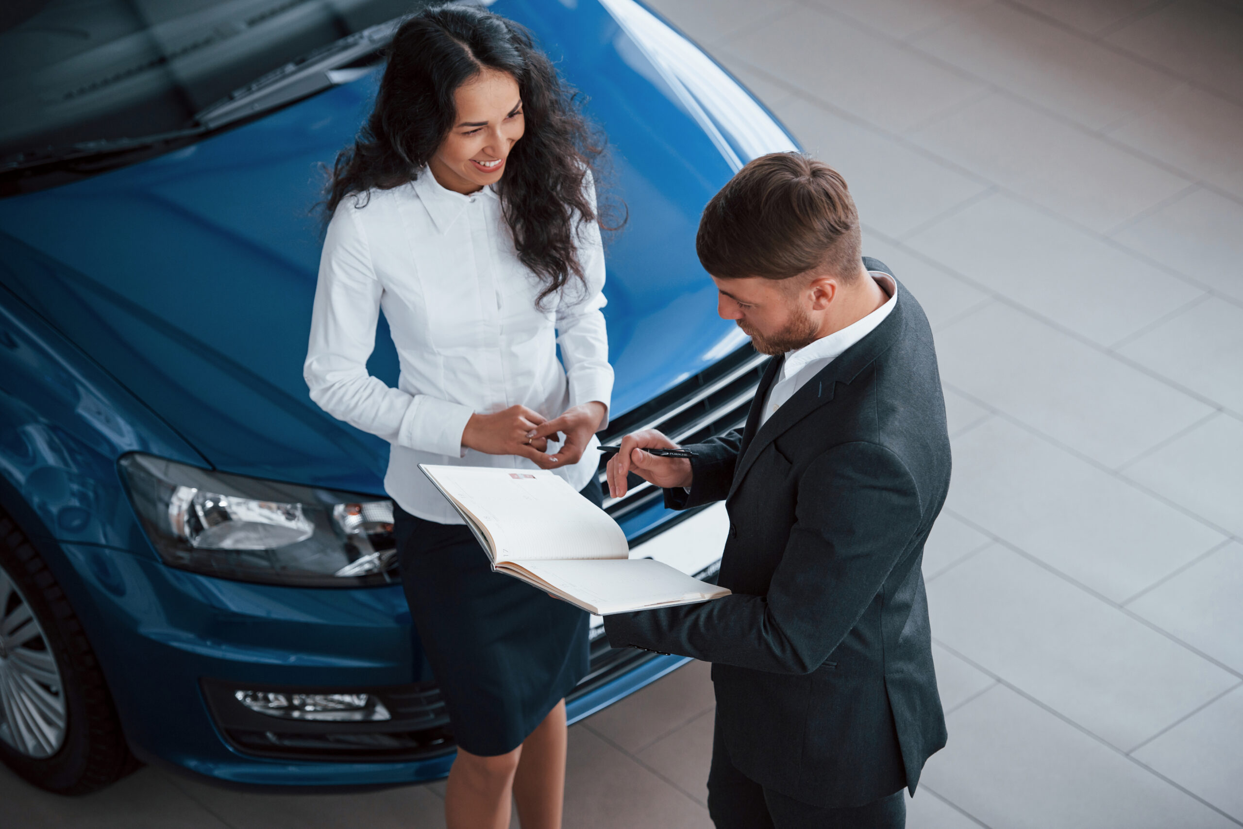 Man and woman in business attire standing by a blue car, reviewing documents.
