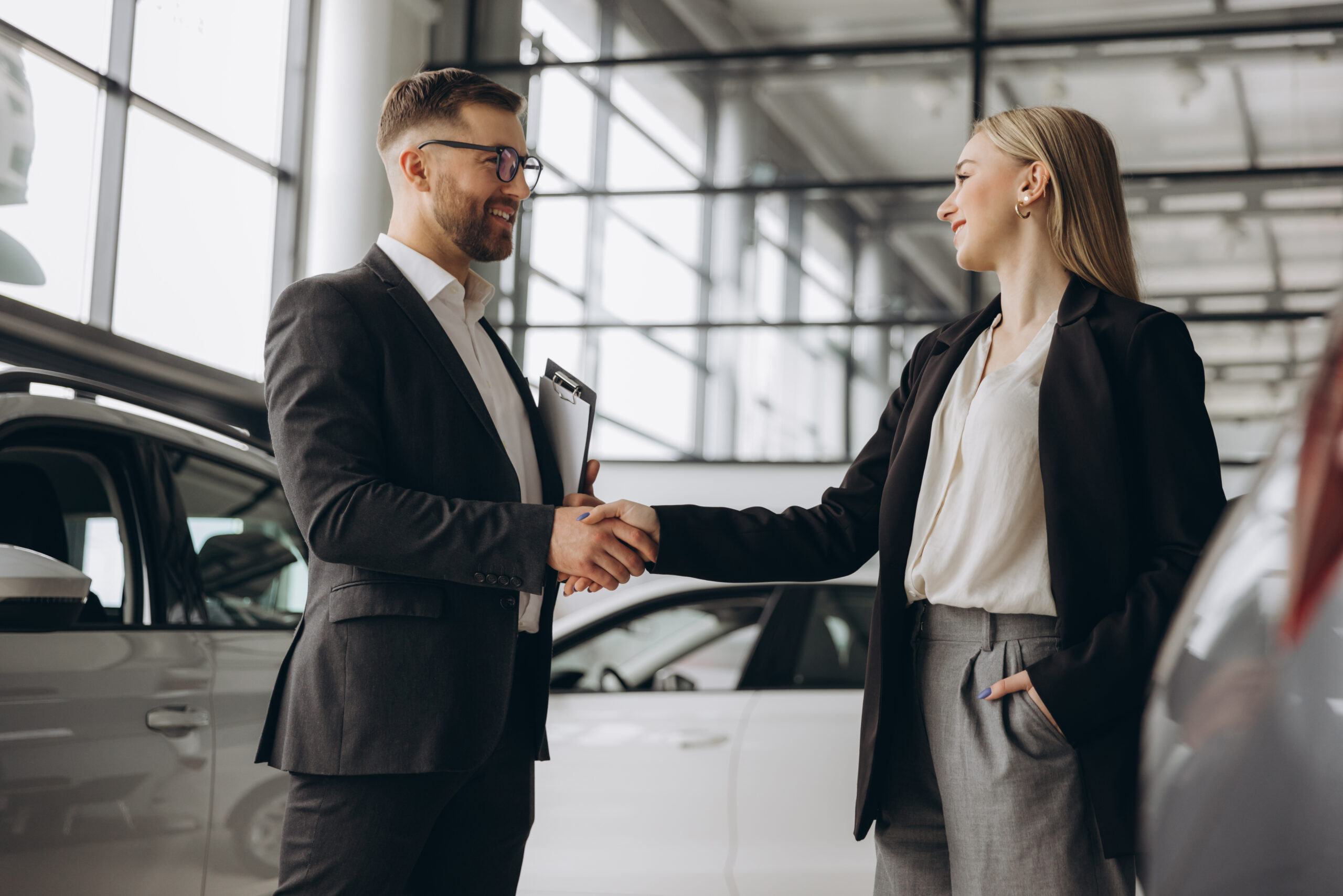 Man and woman shaking hands in a car dealership with a clipboard.